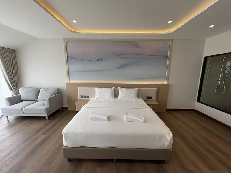 Bedroom with a large bed, a grey sofa, a wooden floor and a pale painted panel above the bed