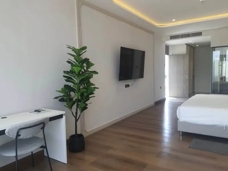 White desk with a chair, a potted plant and a wall-mounted TV, with a bed in the background