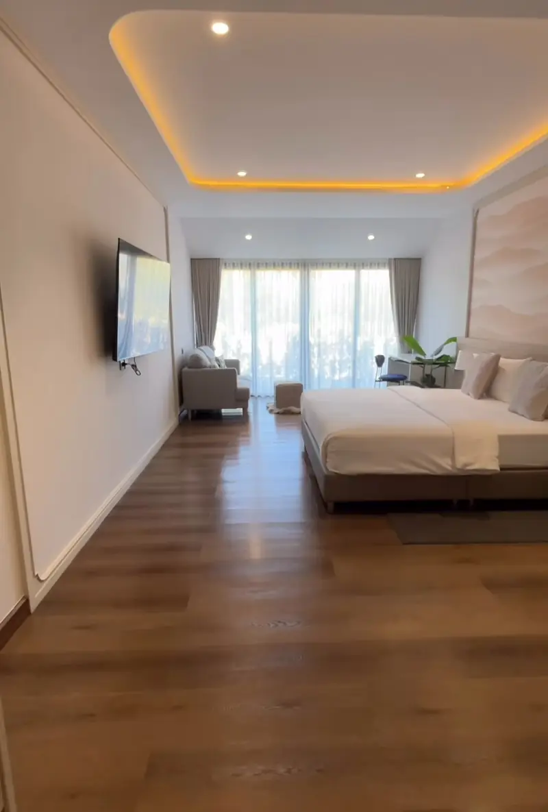 Long view past a wall-mounted TV and an armchair to a bed and tall windows